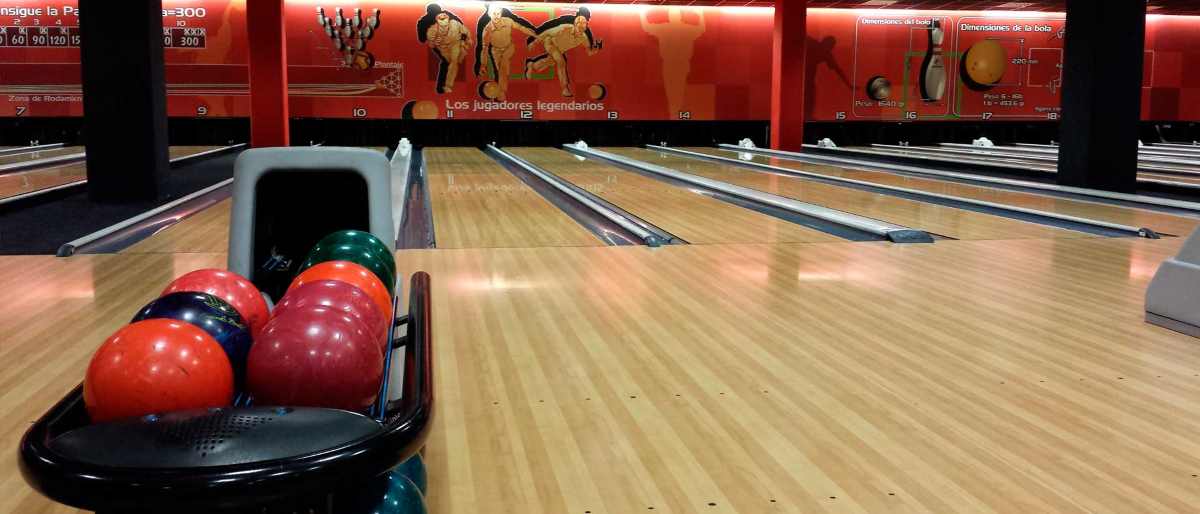 A rack of colorful bowling balls sits in the foreground of a bowling alley with multiple wooden lanes.