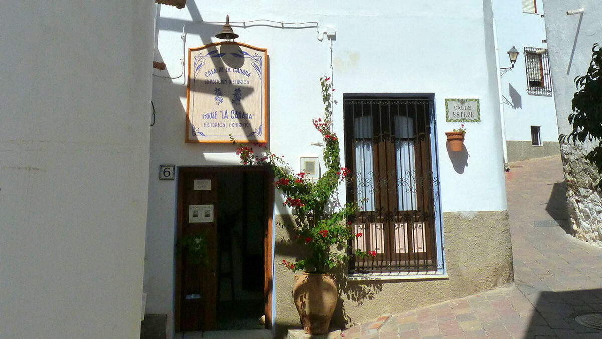 A white building facade features a sign for Casa de la Canana, a doorway, a barred window, and a potted flowering plant.