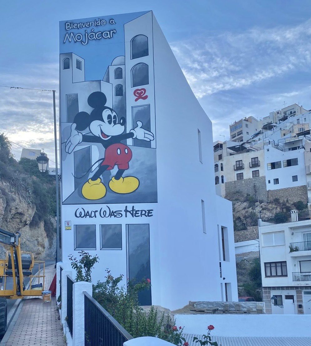 A mural of a cartoon mouse on the side of a white building in a hillside town with text reading Bienvenido a Mojácar.