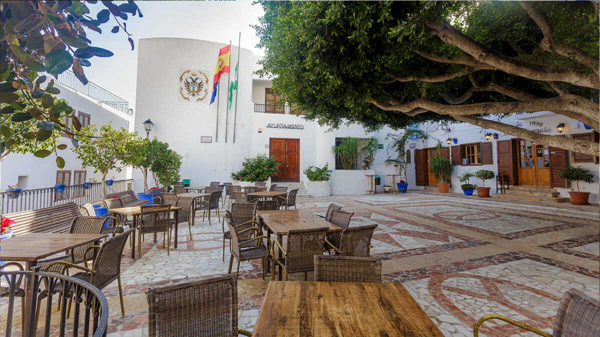 Outdoor tables and chairs are arranged on a tiled patio in front of a white building with a large tree and flags.