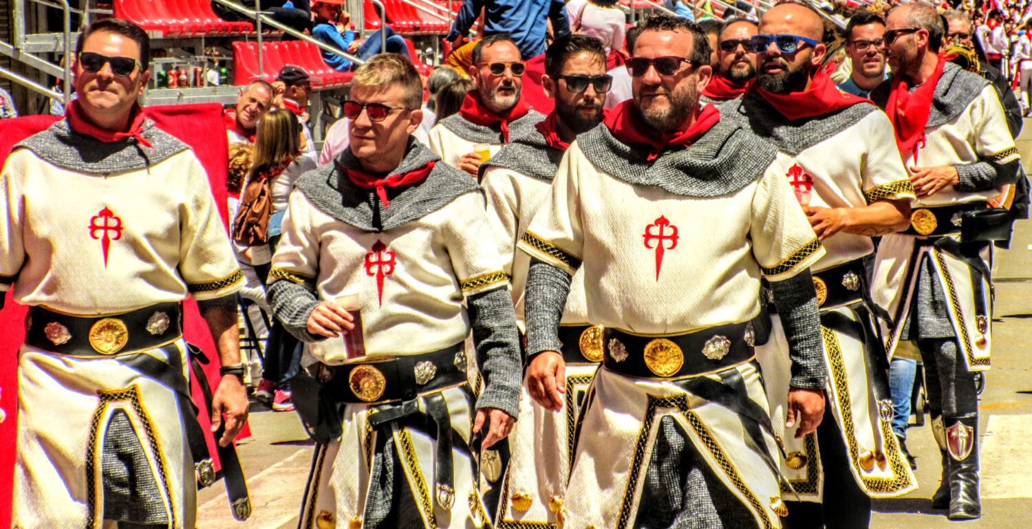 A group of men in medieval-style tunics with red crosses and chainmail hoods walk past red stadium seating.