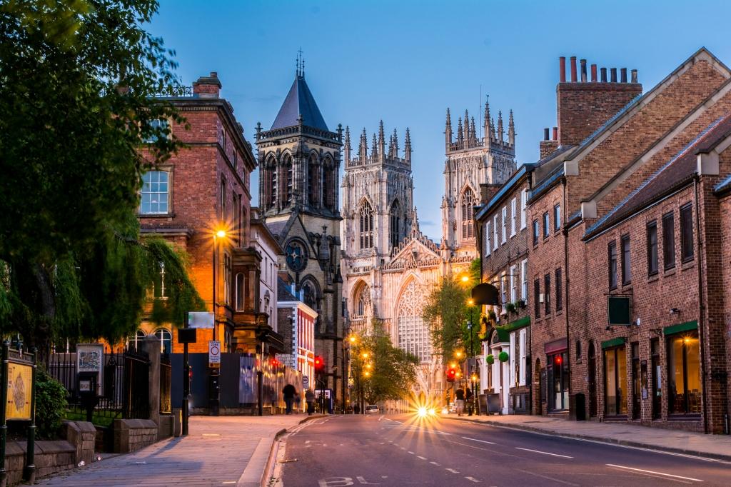 A street leads toward a large, ornate stone cathedral at dusk, flanked by historic brick buildings and glowing streetlights.