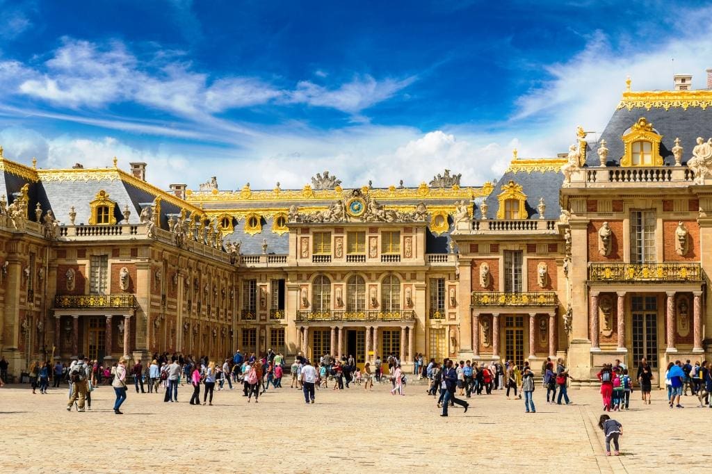 Famous palace Versailles Versailles day trip from Paris. The stunningly decorated with statues and gold trim on the Palace of Versailles with dozens of tourists milling around the front entrance
