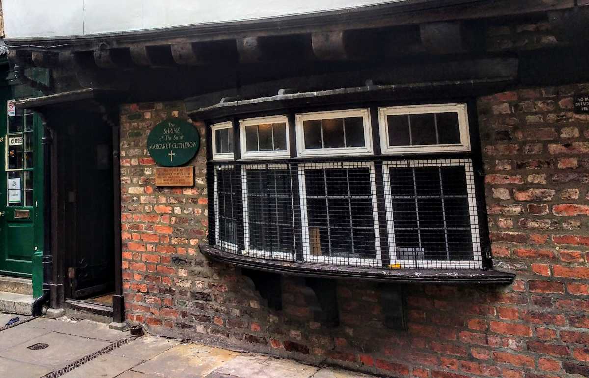 A brick building features a large bay window with a wire mesh screen and a sign for The Shrine of Saint Margaret Clitherow.
