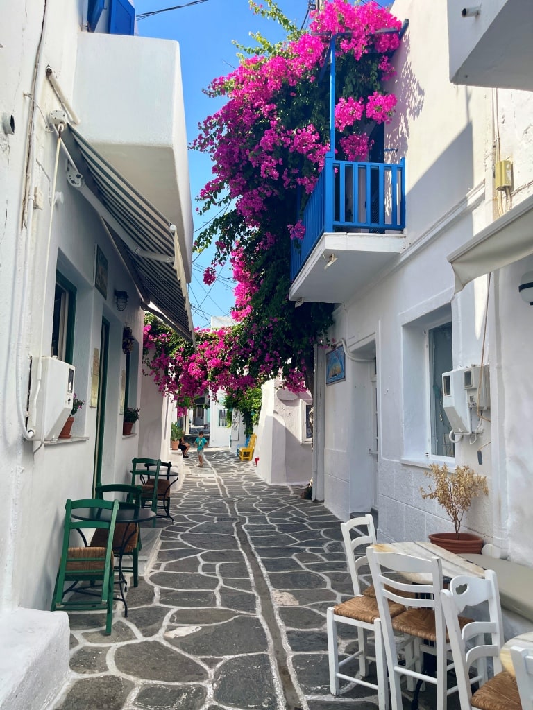 Beautiful whitewashed houses on the island of Paros Greece. Fuschia pink flowers tumble over a blue railing on the 2nd floor of the house. Is Paros worth visiting?