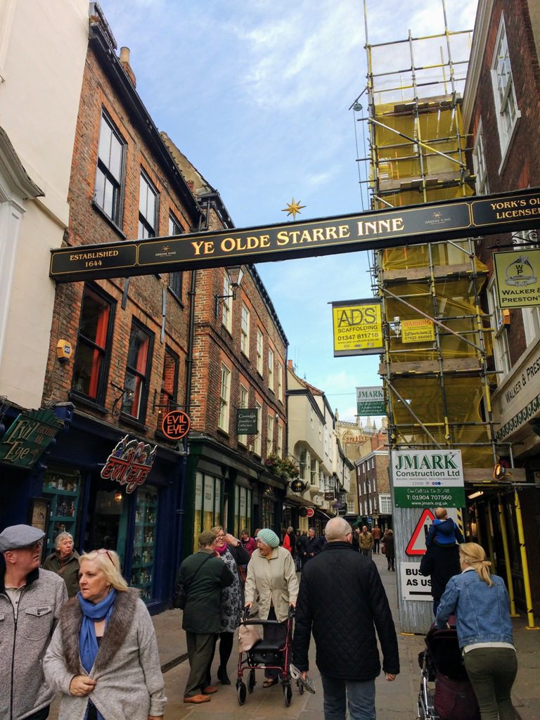 People walk along a narrow street beneath a sign for Ye Olde Starre Inne, with scaffolding visible on a brick building.