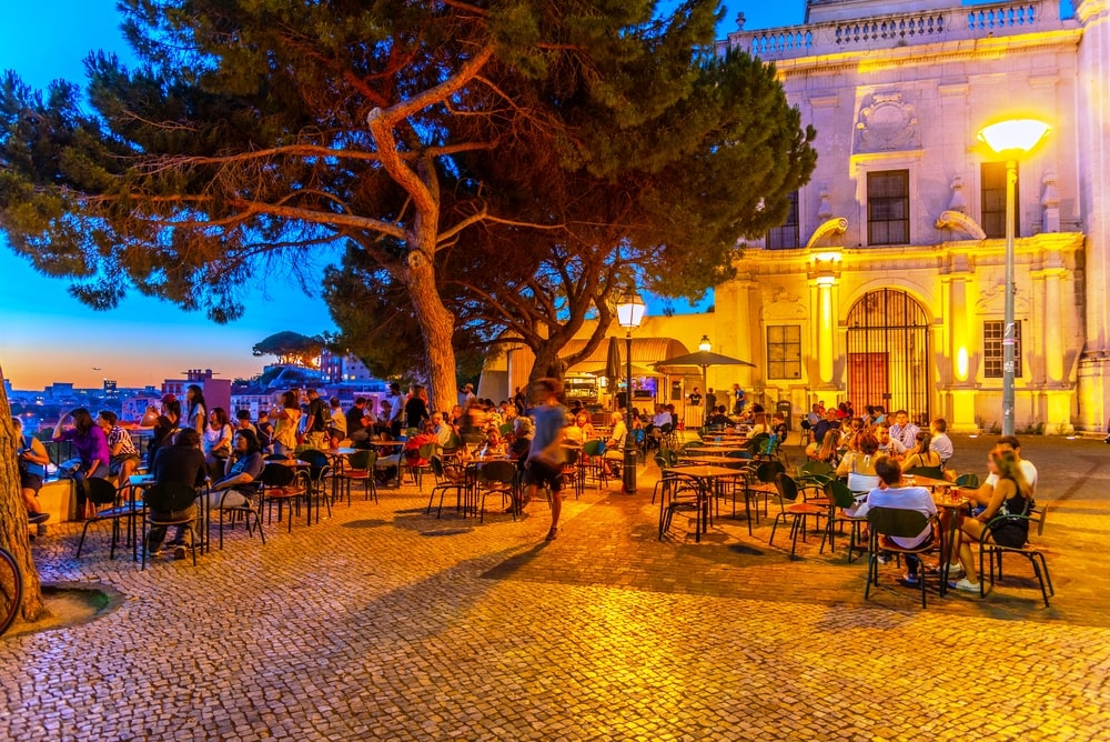 People are enjoying sunset at the Graca viewpoint in Lisbon, Portugal A group of people sitting at tables in a square at dusk.