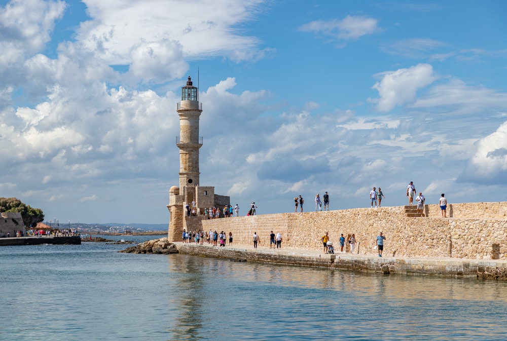 A stone wall with a light tower in Chania Crete, the ancient stone seawall was built by the Egyptians and there are tourists walking along the top of the wall and on the lower wall by the sea of Crete