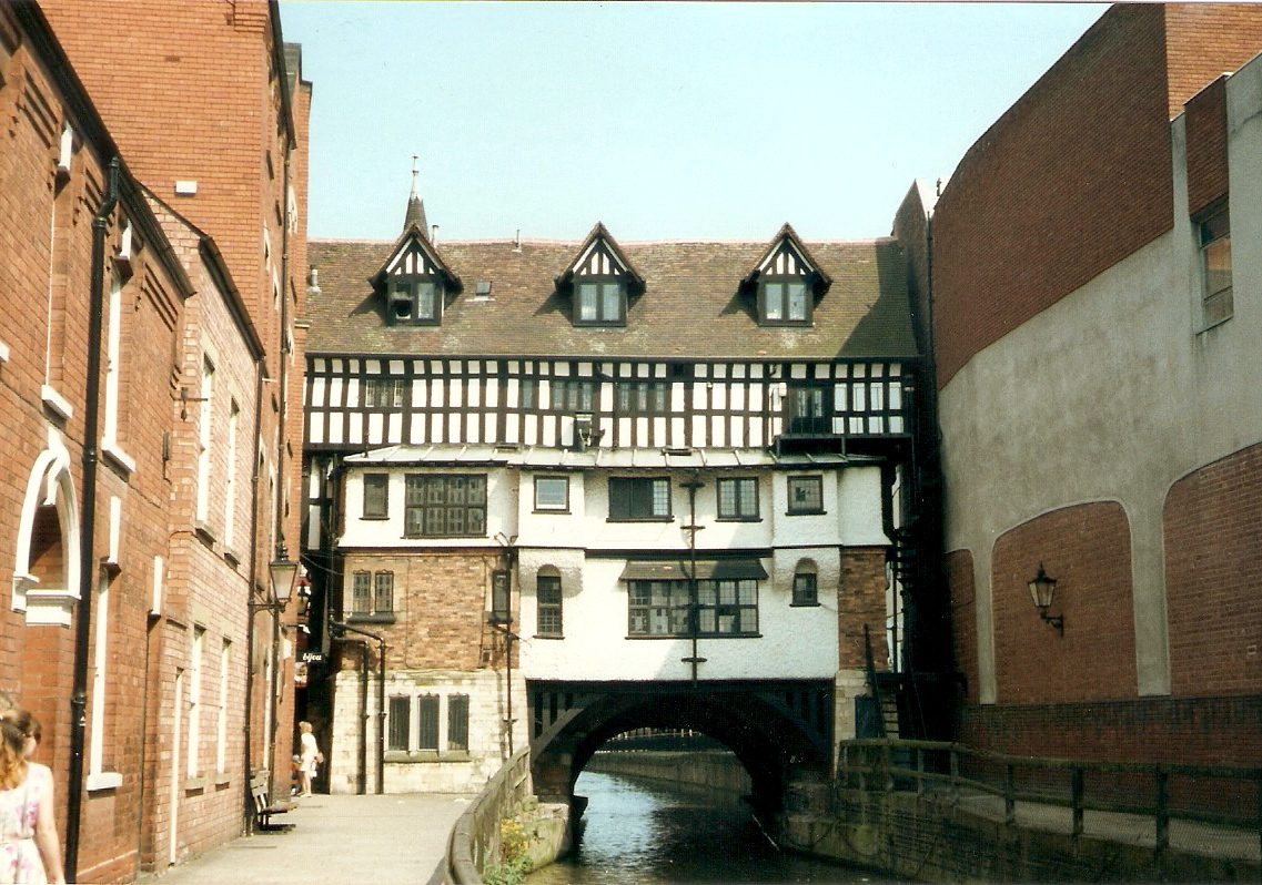 A half-timbered building spans a narrow canal between two brick buildings in an urban setting.