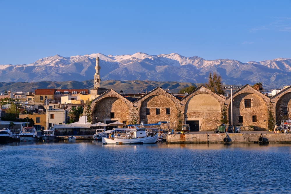 Fishing boats anchored by piers of Old Venetian shipyards known as Arsenali Veneziani or Neoria. Bell tower and minaret of church of Agios Nikolaos and distant Cretan mountain covered by snow in background. Chania, Crete, Greece.