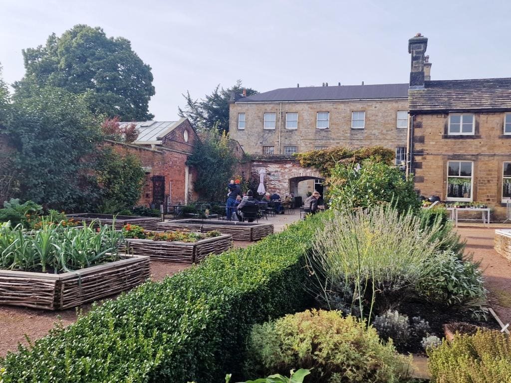 An English manor garden in front of a building with people walking around.