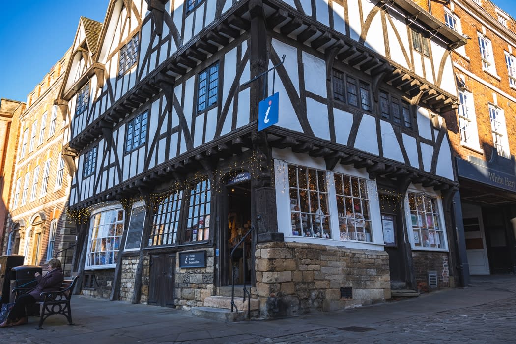 A timber-framed building with a visitor information sign stands on a street corner with a person sitting on a nearby bench.
