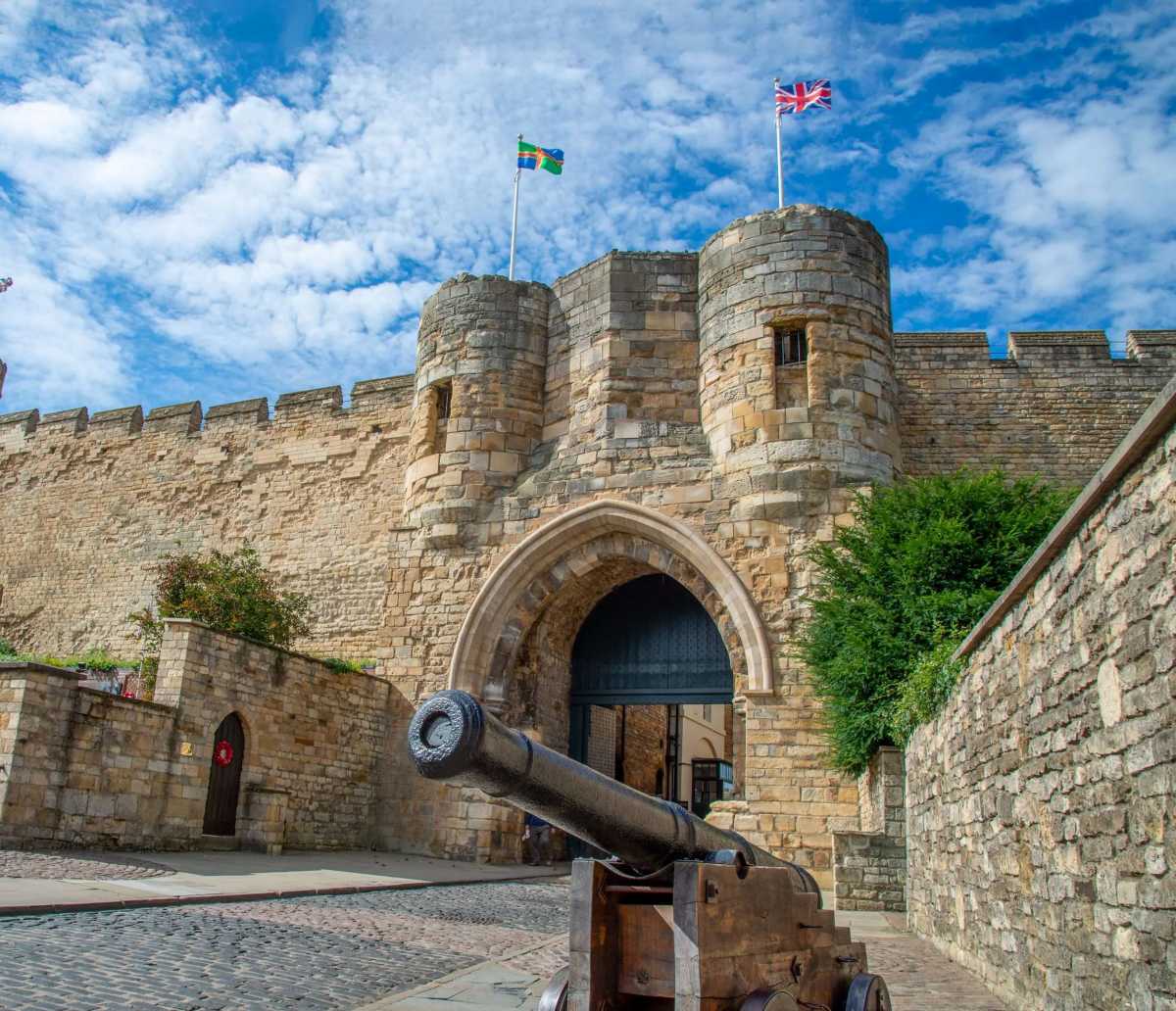 A stone gatehouse with two towers stands behind a cannon on a cobblestone path under a blue sky with clouds.