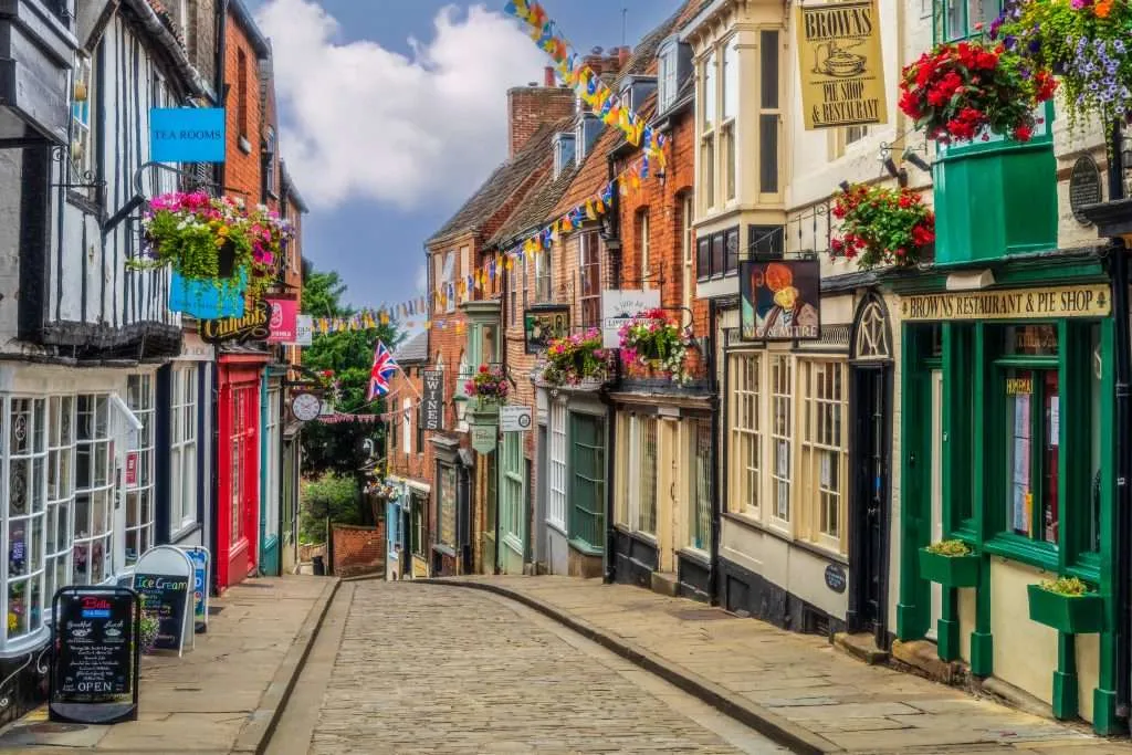 A narrow, cobblestone street is lined with historic brick buildings, storefronts, hanging flower baskets, and bunting.