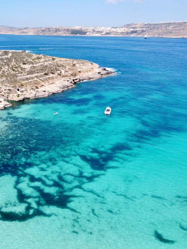 Aerial view of a clear turquoise sea with a boat and swimmer near a rocky coastline, showcasing things to do in Malta.