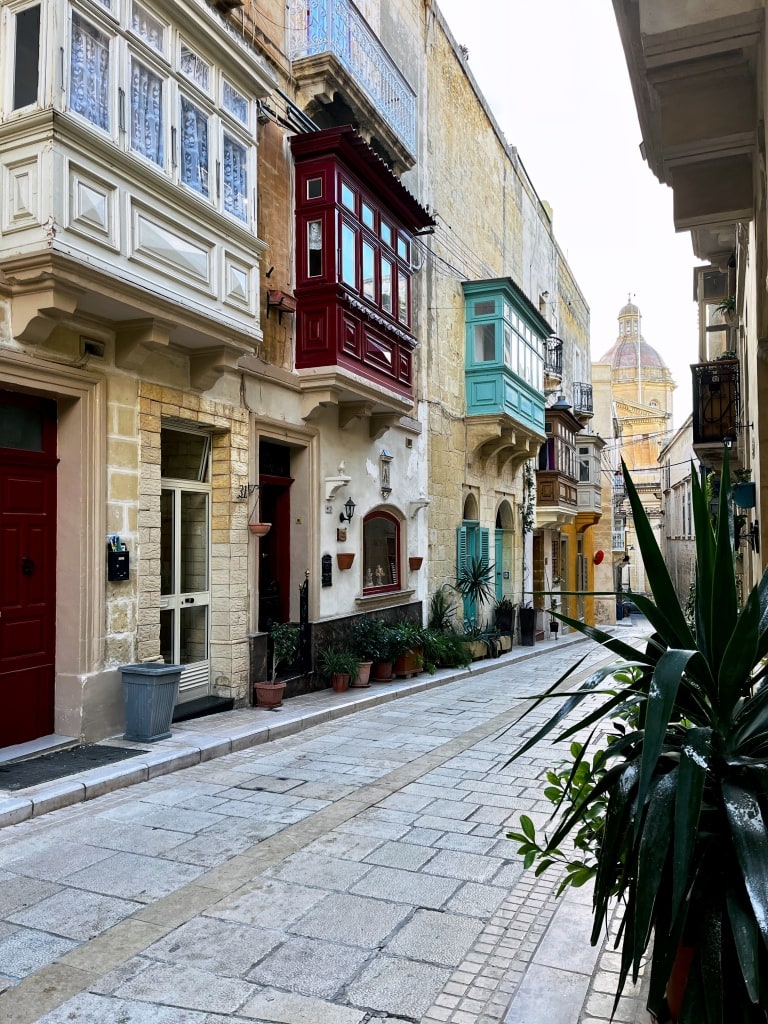 Traditional maltese balconies line a cobblestone street in a quiet, historical urban area.