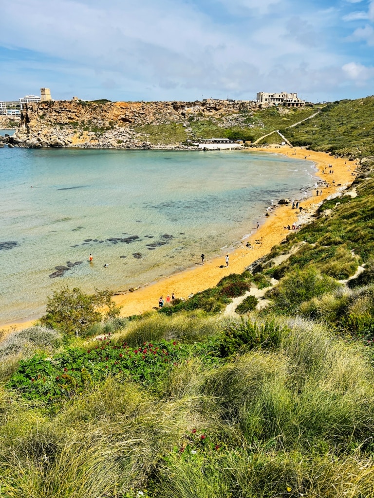 A scenic coastal landscape with a sandy beach, clear shallow waters, and a hillside with greenery under a partly cloudy sky.