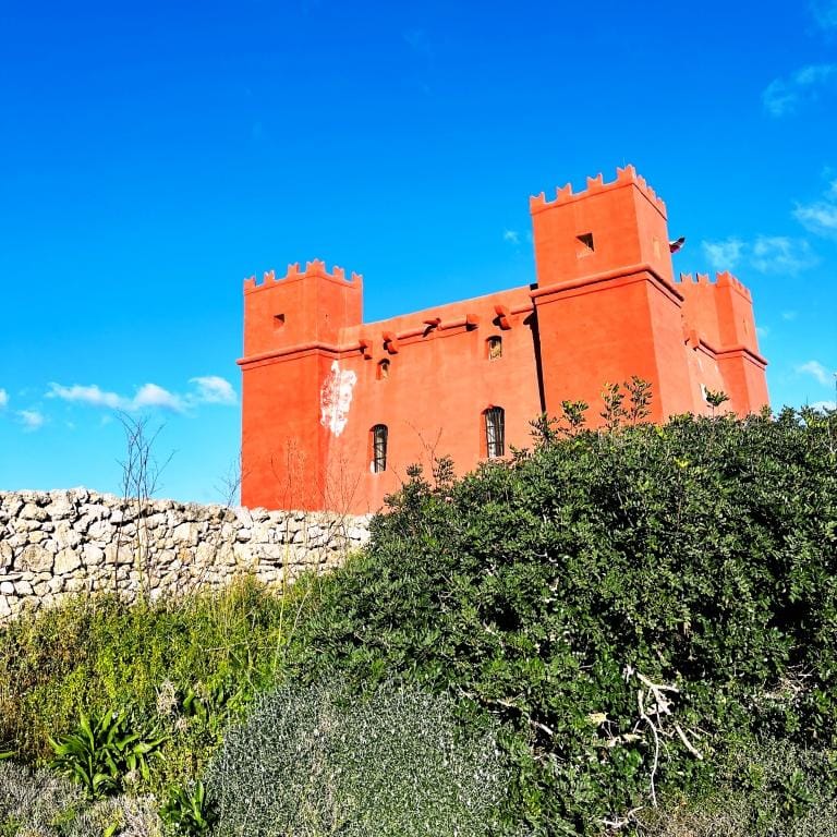 A vibrant orange castle with crenellated towers against a blue sky, surrounded by greenery and a stone wall.