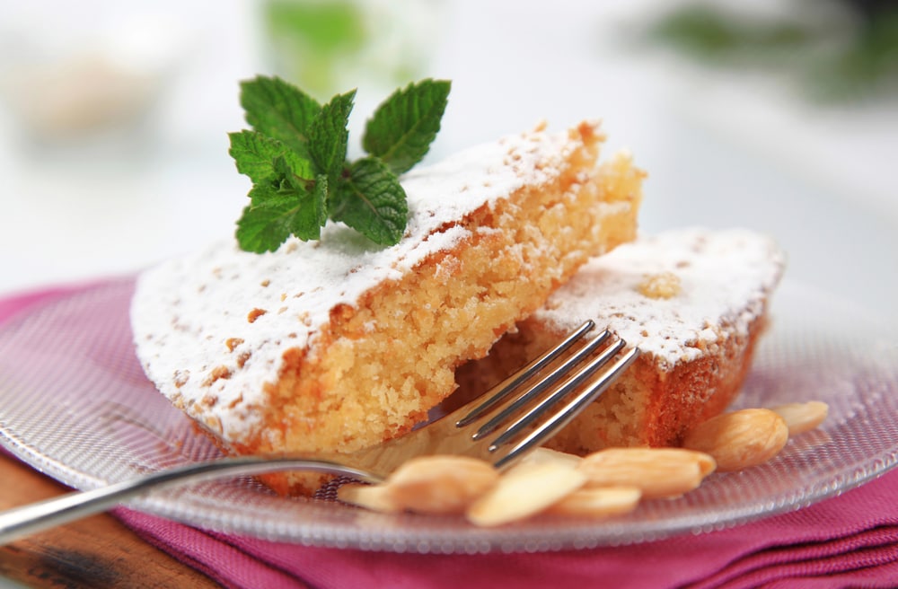 Norwegian Almond cake. A piece of cake on a plate with a fork, with powedered sugar on top and a mint leaf and almonds decorating the plate
