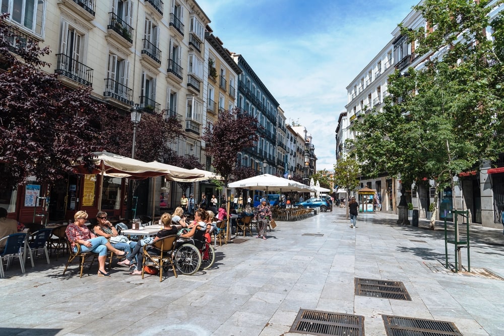 A bustling street scene in Madrid with people sitting at outdoor café tables on a sunny day, surrounded by European-style buildings.