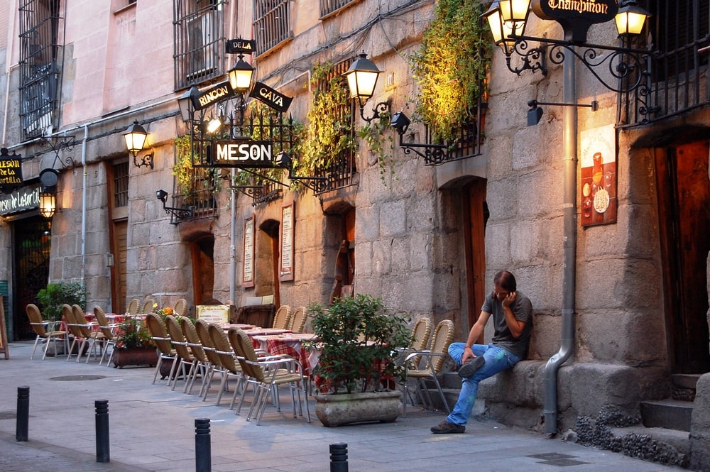 A person sitting on a ledge by a cobblestone street with outdoor restaurant seating and hanging plants in Madrid, enjoying the evening.