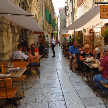 Outdoor dining in a narrow stone-paved alley in Split, Croatia, with customers seated at tables under umbrellas.