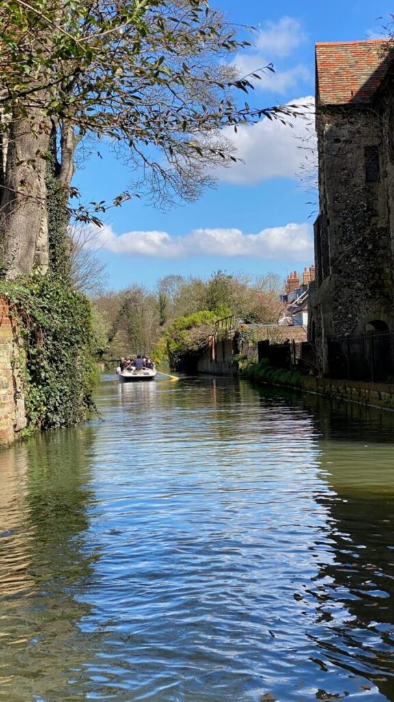 A scenic canal with a Canterbury boat trip, flanked by buildings and greenery under a blue sky with clouds.