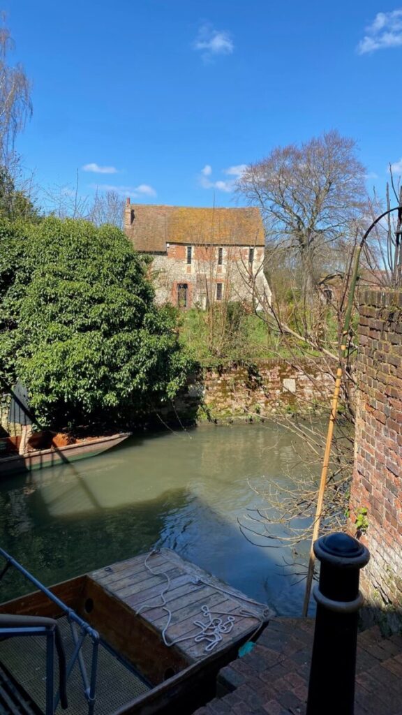 A rustic brick house overlooking a tranquil pond with greenery and a small jetty, perfect for Canterbury boat trips.