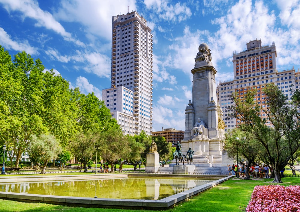 Urban park with a monument and reflective pond in Madrid, flanked by tall buildings under a blue sky.
