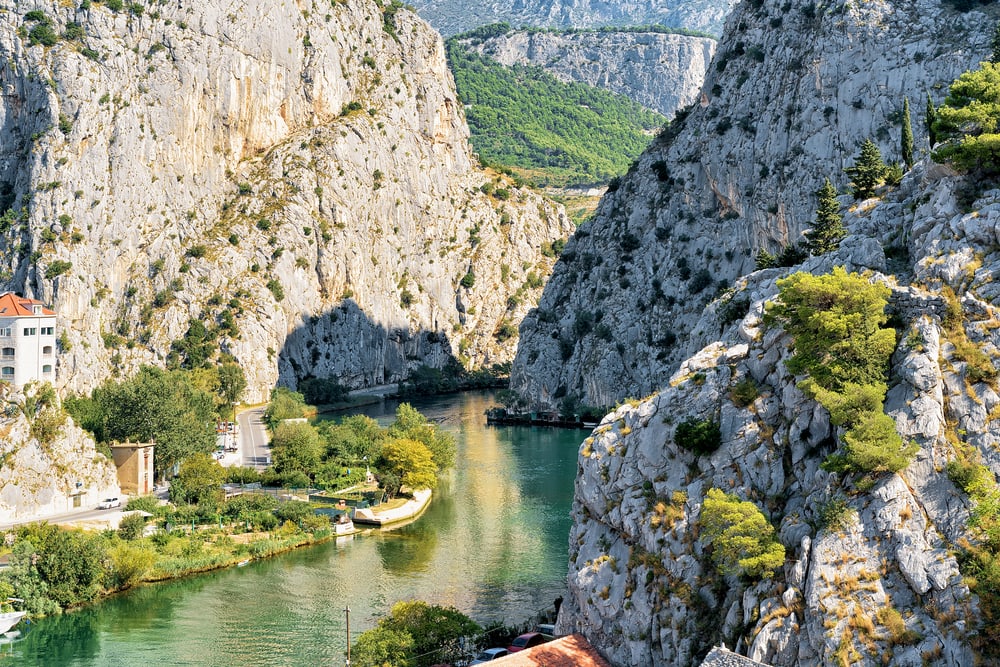 Gorge and Cetina River in Omis, Dalmatia, Croatia. A serene river flowing through a rugged mountain gorge with sparse vegetation and buildings along the banks, offering captivating things to do in Split, Croatia.
