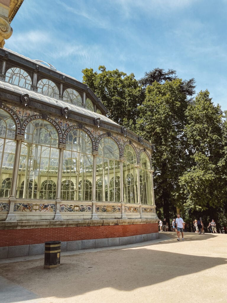 Ornate glass and metal greenhouse structure with visitors enjoying one day in Madrid, in a sunny park setting.