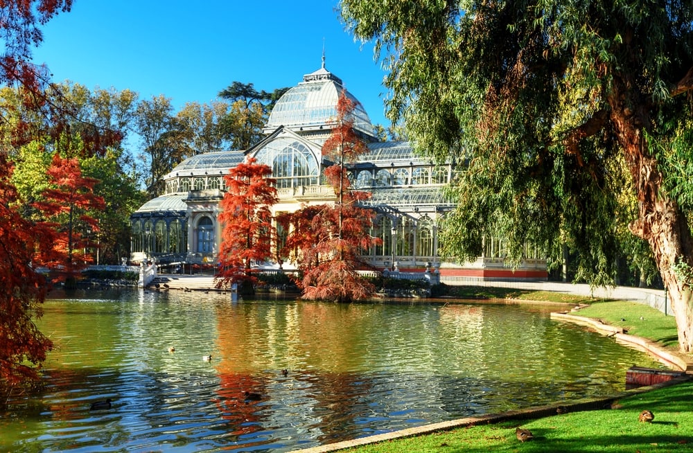 Crystal Palace in Retiro Park, Madrid, surrounded by autumn foliage and reflecting in a tranquil pond, is a must-visit on one day in Madrid.