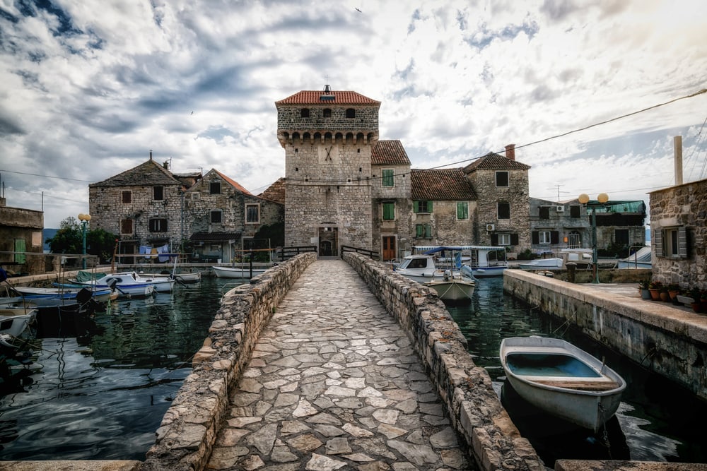 Kastel Gomilica is the filming location of Game of Thrones TV series in Croatia. Kastel Gomilica is one of seven settlement of town Kastela in Croatia. Stone pathway leading to an old fortified building by a serene marina with moored boats under a dramatic sky, showcasing things to do in Split, Croatia.