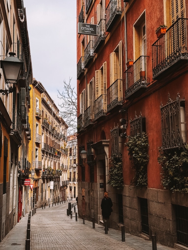 A person walking down a quiet, narrow cobblestone street lined with traditional European buildings, experiencing one day in Madrid.