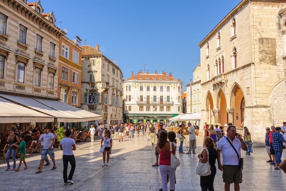 Tourists and locals stroll through a bustling European city square in Split, Croatia, lined with historic buildings and outdoor cafes.