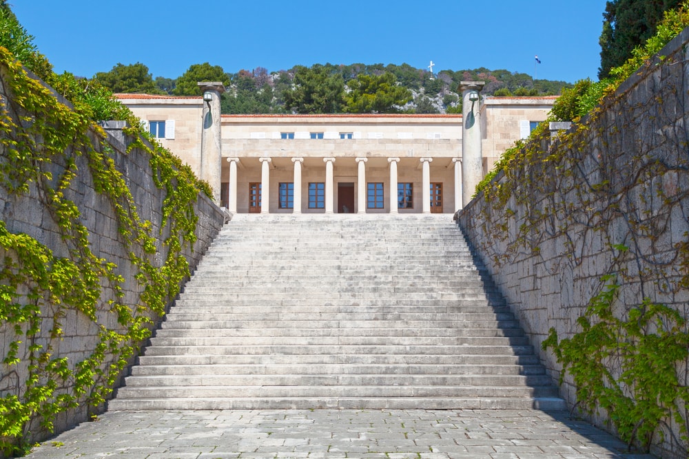Stone staircase leading up to a classical building with columns in Split, Croatia, surrounded by green foliage under a clear blue sky. The Mestrovic Gallery (Croatian: Galerija Meštrović) is the former residence & studio of sculptor Ivan Meštrović. It is now an art museum displaying his works.