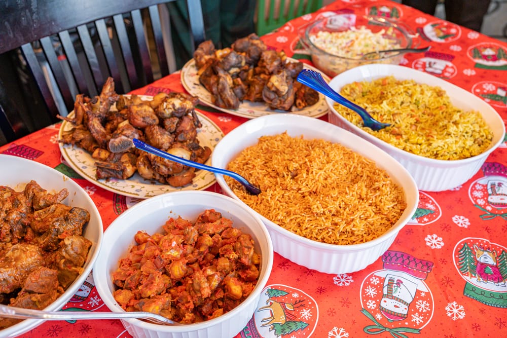 Variety of West African dishes on a festive tablecloth including grilled chicken, jollof rice, and a tomato-based stew.