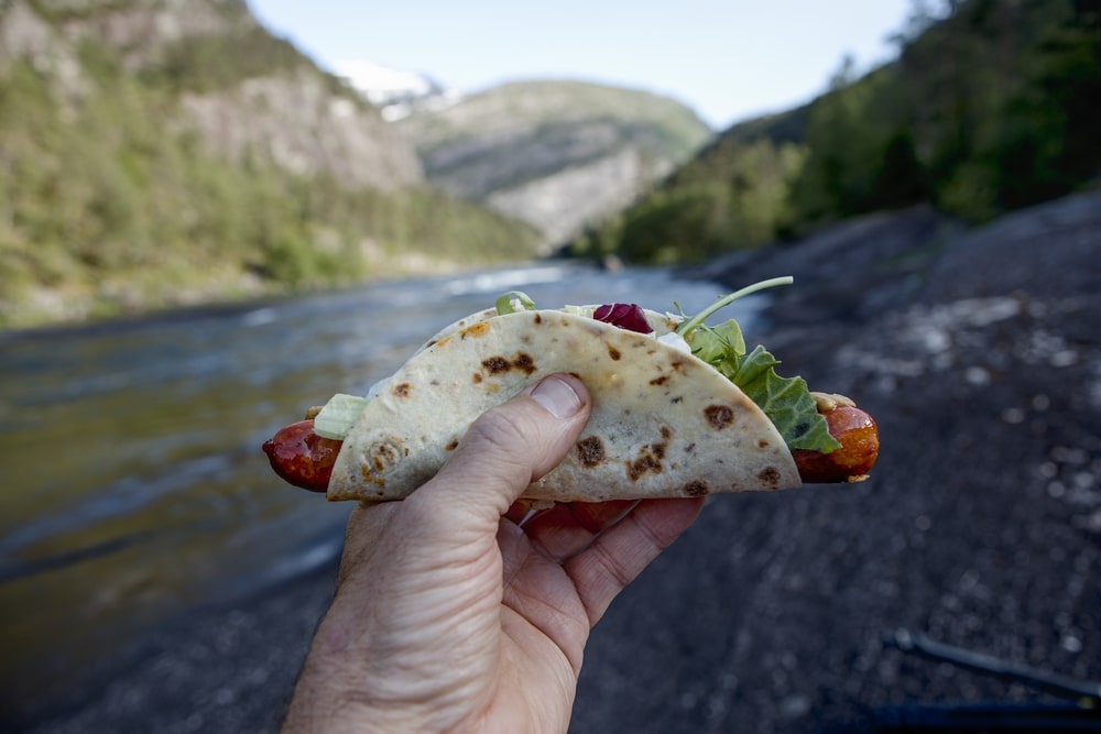 A person holding a hot dog wrapped in Lefse, a popular food in Norway, in front of a river.