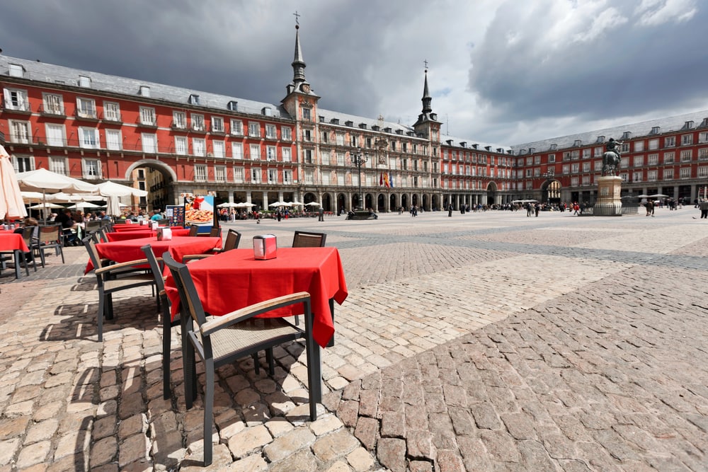 Outdoor dining area with red tablecloths in a historic European square under a cloudy sky, capturing the essence of one day in Madrid.