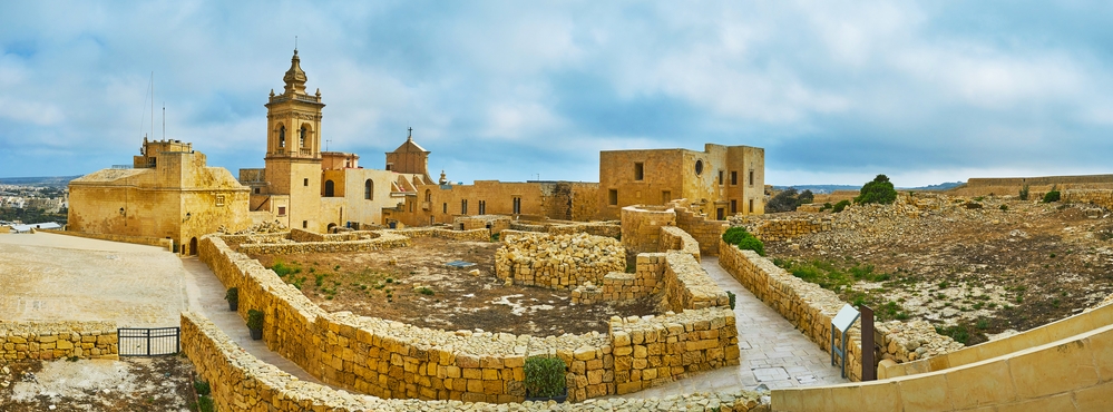 Panorama of the Rabat Citadel with foundations of ruined buildings, St John Cavalier and Assumption Cathedral, Victoria, Gozo Island, Malta.. Explore things to do in Malta, including visiting the panoramic view of the historic citadel with limestone architecture under a cloudy sky.