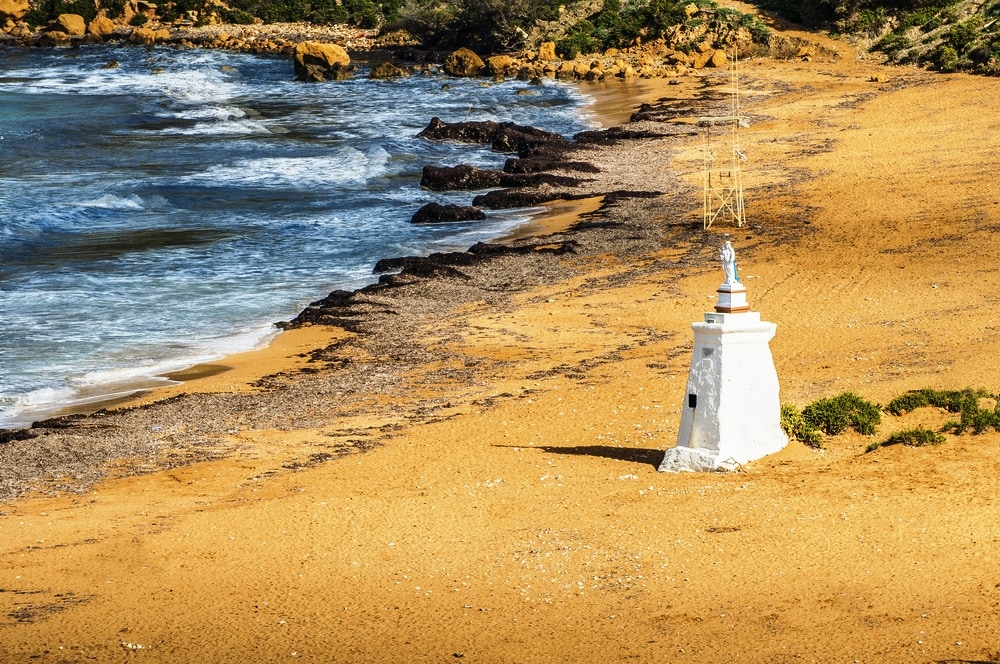 Ramla Beach on the northern side of Gozo, Malta. A small white lighthouse stands on a sandy beach with rocky outcrops along the shoreline and waves lapping at the shore, offering picturesque views for those exploring things to do in Malta.