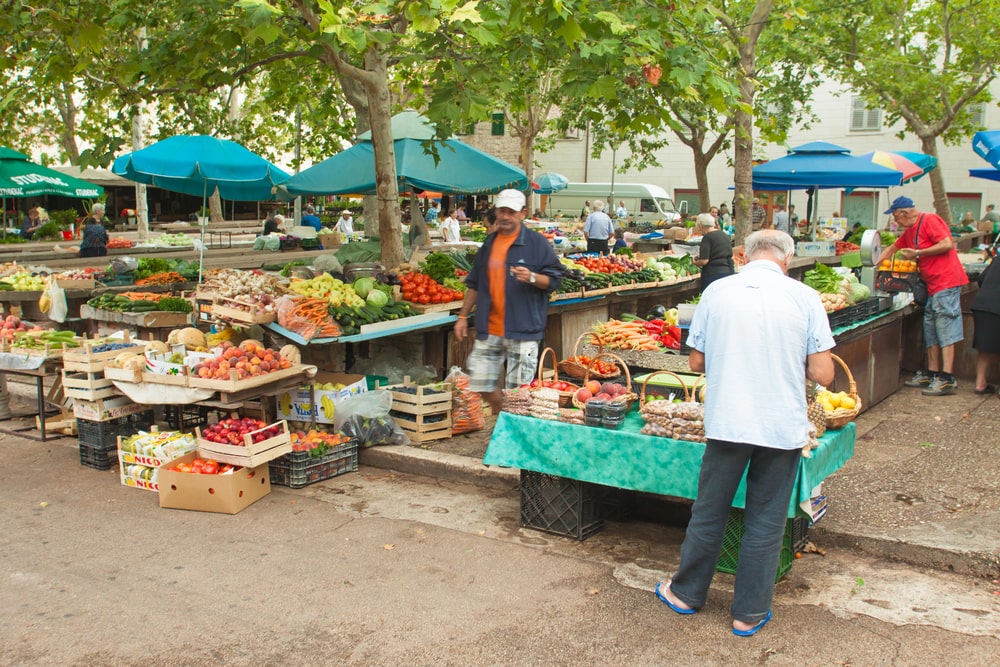 Outdoor market stalls displaying fresh produce under blue umbrellas with shoppers and vendors, offering a glimpse into the things to do in Split Croatia.