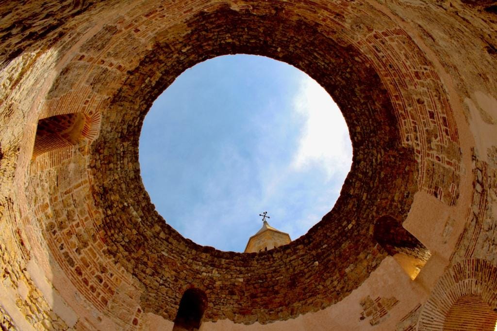 Upward view of a round ancient structure in Split Croatia, opening to a blue sky with a cross silhouetted against the sky.
