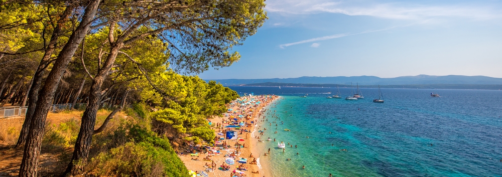 Famous Zlatni rat beach in Bol, Island Brac, Croatia. Panoramic view of a busy beach in Split, Croatia, with visitors enjoying the sun, lined by trees on one side and clear blue water with boats on the other.