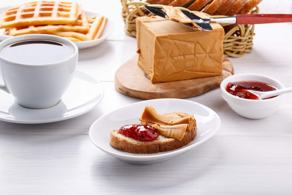 A cup of coffee on a table next to a plate of waffles and jam, emulating the cozy food experience in Norway. Norwegian brunost on white table. Breakfast with Scandinavian brown cheese, bread and coffee.