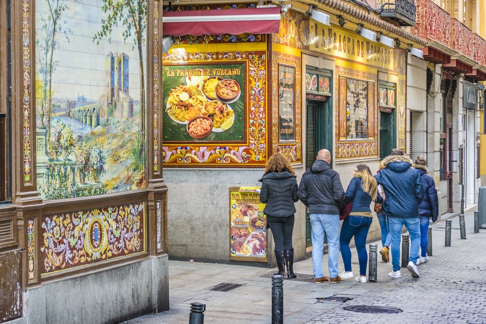 Pedestrians pass by a colorful, tiled restaurant facade in a quaint Madrid street.