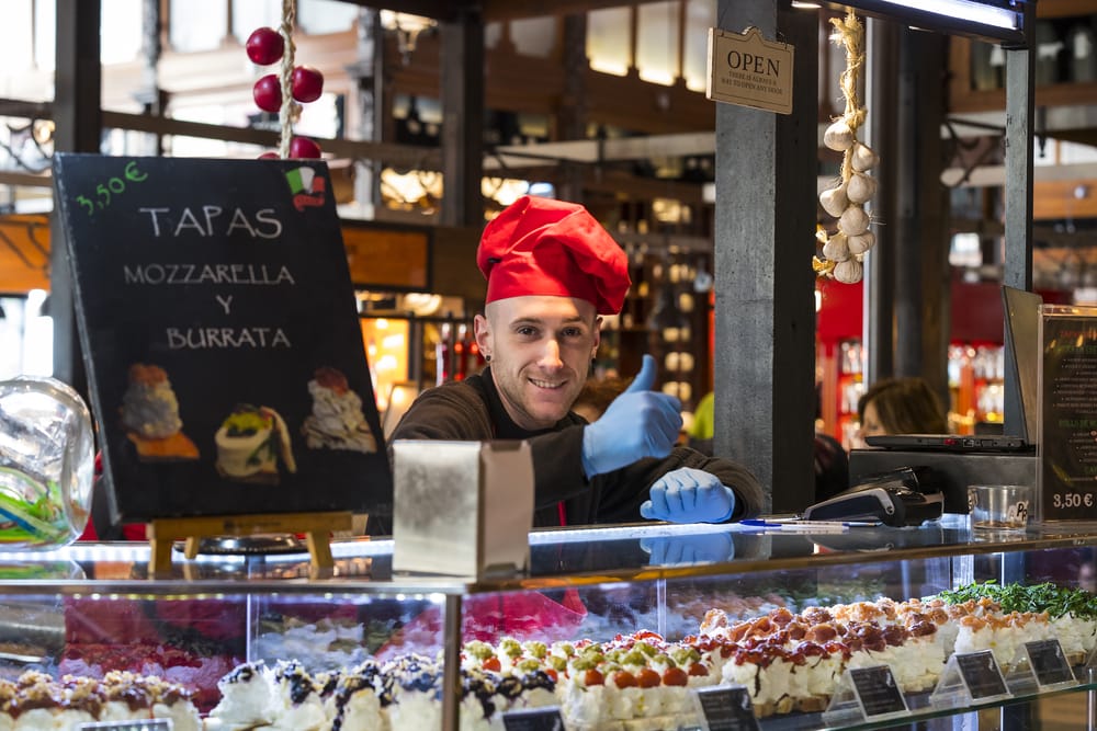 Chef wearing a red hat smiles from behind a food counter with an assortment of tapas, one day in Madrid, next to a chalkboard sign displaying prices.