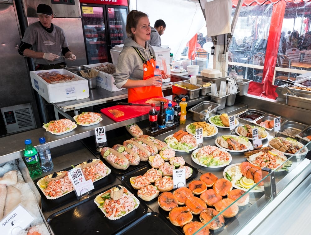 Various seafood on the shelves of the fish market in Bergen in Norway. A woman standing behind a counter serving traditional food in Norway. On the counter are platters of fishes including lobster rolls, smoked salmon plates, shrimp and prawn salads and Norwegian smoked salmon on bagels. 