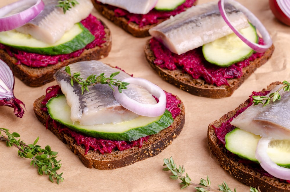 A plate of traditional food in Norway with fish, onions, and beets on it. The bread is openfaced dark rye bread with a beetroot saled topped with cucumber and smoked herring, a spring of thyme and raw onions 