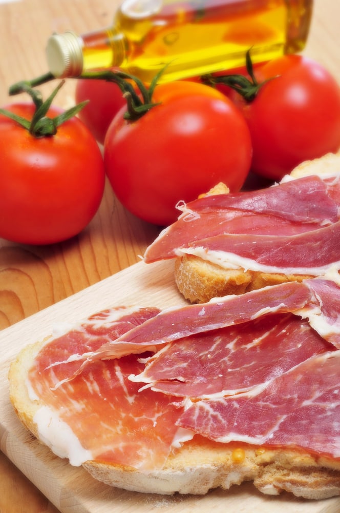 Slices of prosciutto on bread with tomatoes and olive oil in the background, reminiscent of one day in Madrid.